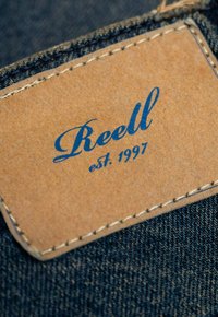 Braunes Lederetikett mit blauer Schrift "Reell est. 1997", auf dunkelblauem Denim-Stoff angenäht, mit sichtbaren Nähten entlang der Kanten.