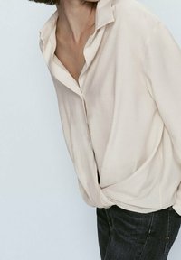 Chemise beige clair à manches longues avec un col, coupe ample, partiellement rentrée dans un jean en denim foncé, présentant une texture de tissu douce.