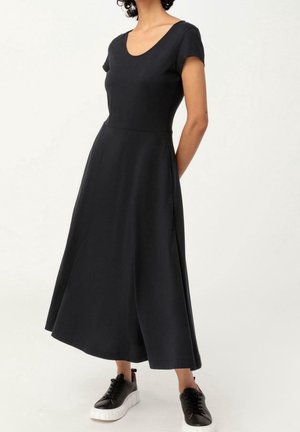 Femme portant une robe midi noire à manches courtes associée à des baskets montantes noires sur un fond blanc uni.