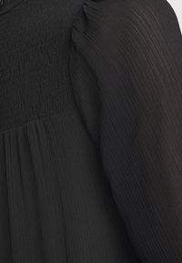 Tissu noir avec partie supérieure froncée smockée et détail de manche semi-transparent finement plissé.