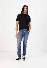 Black short-sleeve t-shirt, blue denim jeans, and black sneakers with white soles. Model stands in a neutral background, hands in pockets.