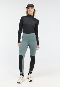 Zwart langslang top met halve rits, gecombineerd met aansluitende teal en zwarte leggings. Het model draagt beige sneakers en een zwarte pet.