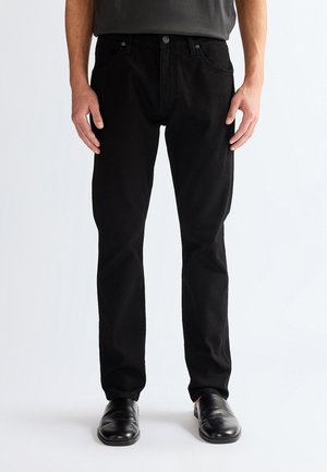 Muškarac u crnim trapericama ravnih nogavica, crnim kožnim cipelama i tamnosivoj košulji, stoji ispred bijele pozadine.