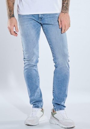 Mann trägt hellblaue Slim-Fit-Jeans und weiße Sneaker, steht vor einem einfarbigen hellen Hintergrund.