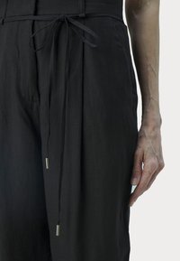 Svart linneoverall med knytmidja, som har sidofickor och en mjuk yta. Accentdetaljer inkluderar tunna, metalliska snoddar.