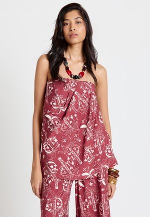 Femme portant un haut rouge sans bretelles et un pantalon assorti avec imprimé blanc de palmier tropical, accessoirisée avec un collier de perles rouge et noir épaisses et des bracelets.