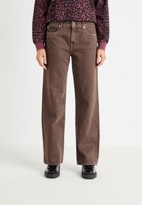 Barna, bővített lábú farmernadrág, gombos záródással, öt zsebbel és finom fakulással. Fekete cipővel párosítva.