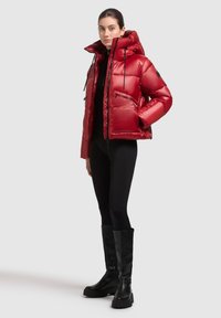 Chaqueta acolchada roja con un collar alto, bolsillos con cremallera y una textura brillante. Combinada con pantalones negros ajustados y botas altas negras.