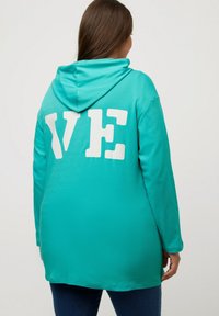 Sudadera con capucha turquesa de manga larga, con grandes letras blancas "VE" en la parte de atrás. Textura suave, corte relajado y puños acanalados.