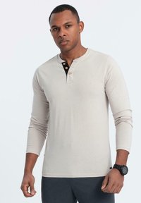 Man som bär en beige långärmad henley-tröja med en svart innerknäppning och mörka byxor, står mot en enkel ljus bakgrund.