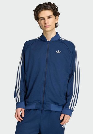 Junger Mann, der eine blaue Adidas Trainingsjacke und passende Hose mit weißen Streifen trägt, steht vor einem schlichten hellen Hintergrund.