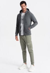 Grauer Zip-Up-Hoodie über einem weißen Grafik-T-Shirt, kombiniert mit olivfarbenen Cargo-Hosen und weißen Sneakers, die Taschen und elastische Bündchen haben.