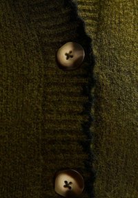 Mörkgrönt stickat tyg med två stora bruna fyrhålsknappar och en svart lurvig kant längs kanten nära knapparna.