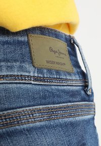 Jeans in denim blu con una texture liscia, dotati di una toppa in pelle marrone scuro etichettata "Pepe Jeans Wiser Wash" e dettagli di cucitura a contrasto.