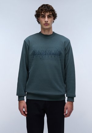 Groene sweatshirt met een ronde hals en geribde manchetten, met een donkerder groene grafische print over de borst. Zwarte broek is zichtbaar eronder.