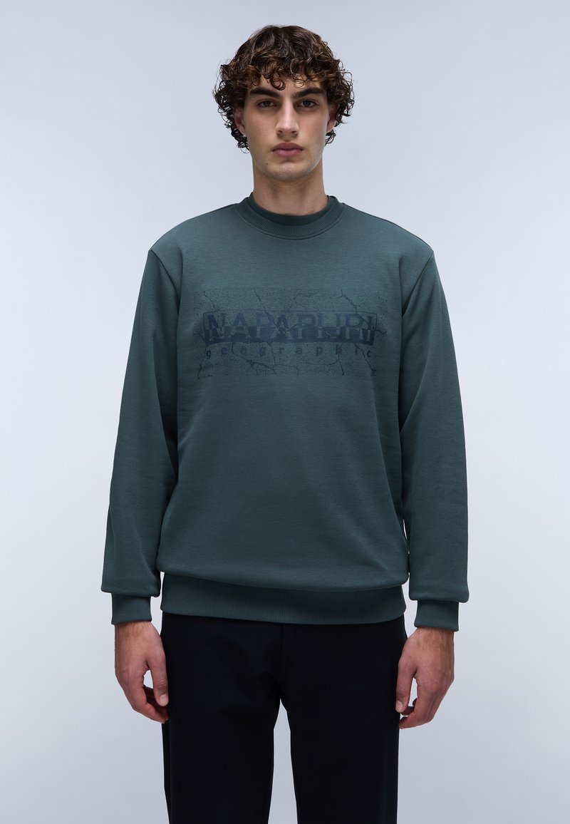 Felpa verde con collo rotondo e polsini a costine, caratterizzata da una stampa grafica verde scuro sul petto. In basso sono visibili pantaloni neri.