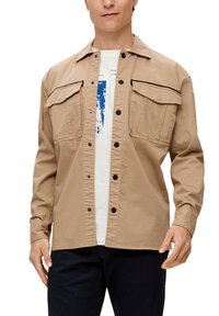 Giacca button-up di color marrone chiaro con due tasche sul petto, dettagli neri e vestibilità rilassata, indossata sopra una camicia bianca. Materiale in cotone, texture liscia.