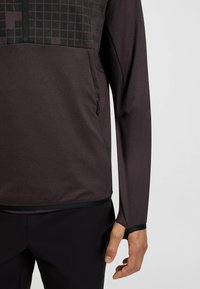 Långärmad pullover i mörklila, med ett texturerat geometriskt mönster på den övre delen och en dragkedjeförsedd ficka fram.