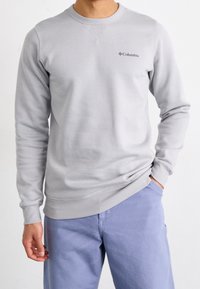 Hombre vestido con un sudadera ligera gris de Columbia y pantalones claros azules, con la mano en el bolsillo, de pie contra un fondo blanco liso.