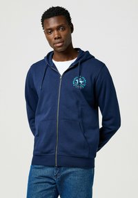 Felpa con zip di colore navy realizzata in misto cotone, dotata di tasca a canguro, polsini a costine e logo ricamato in turchese sul petto.