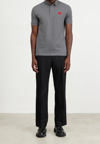 Camisa polo gris de manga corta y cuello, con un parche de logo rojo en el lado izquierdo del pecho. Combinada con pantalones negros y zapatos negros.