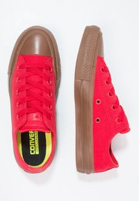 Zapatillas de lona rojas con suelas de goma marrón, puntera redonda y seis ojetes. En el interior cuentan con una plantilla negra y amarilla. Textura suave.