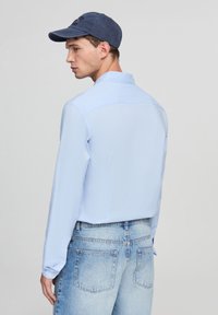 Camicia a maniche lunghe azzurro chiaro con colletto, texture liscia, abbinata a jeans in denim azzurro chiaro. La persona indossa un cappellino blu scuro.