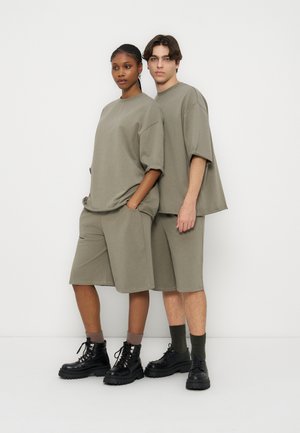 Zwei Models stehen nebeneinander und tragen passende übergroße olivgrüne T-Shirts, lange Shorts, dunkle Socken und schwarze klobige Stiefel.