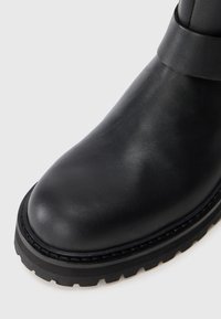 Bota de cuero negro con punta redonda, suela gruesa con dibujo y ribete cosido, fotografiada sobre un fondo blanco.