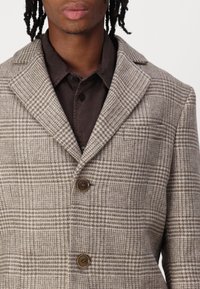 Blazer de cuadros marrón y beige con solapa de muesca, tres botones y tela texturada. Lleva puesto sobre una camisa oscura con botones.