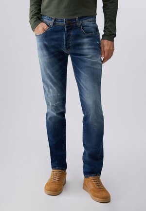 Muž stojící, oblečený v vybledlých modrých džínách s mírným opotřebením a v béžových semišových teniskách, ruce volně podél těla na jednobarevném pozadí.