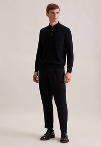 Zwarte gebreide trui met een polokraag, twee knopen en geribbelde manchetten, gecombineerd met donkere taps toelopende broek en zwarte leren schoenen.