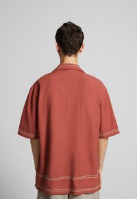 Camisetta a maniche corte di colore ruggine con una vestibilità rilassata, colletto e dettagli in cucitura bianca lungo i bordi e l'orlo.