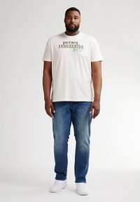 Cremefarvet bomulds-t-shirt med trykt tekst i grøn og sort, kombineret med blå denimjeans og hvide sneakers, båret af en mandlig model.