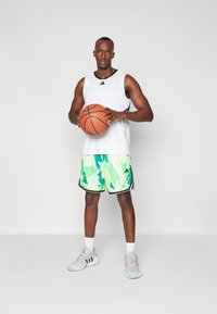 Weißes ärmelloses Trikot, grün und blau gemusterte Shorts, hält einen Basketball. Sneakers mit grauen, blauen und weißen Akzenten.