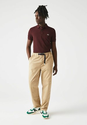 Polo shirt bordeaux a maniche corte, abbinato a pantaloni cargo beige con vita elastica, caratterizzati da una cintura nera e sneaker verdi e bianche.
