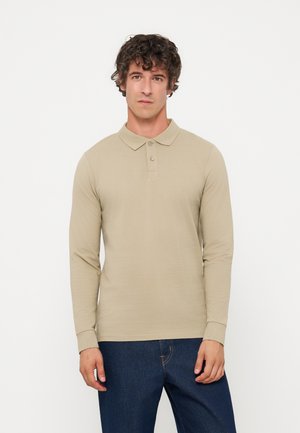 Man met krullend haar draagt een beige poloshirt met lange mouwen en donkerblauwe spijkerbroek, staand tegen een effen witte achtergrond.