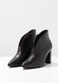 Bottes noires en cuir à la cheville avec bouts pointus, fermetures éclair latérales et talon haut épais. Texture lisse avec un design élégant.