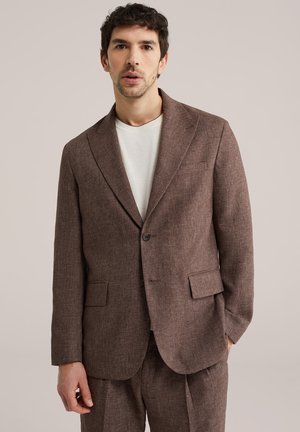 Moški, ki nosi rjavo teksturirano obleko z dvema sprednjima žepoma z zaponkami in belo majico z okroglim izrezom, stoji pred preprostim nevtralnim ozadjem.