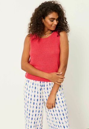 Kobieta o kręconych włosach, ubrana w czerwony top bez rękawów i białe spodnie z niebieskim wzorem liści, stojąca z delikatnie skrzyżowanymi ramionami.