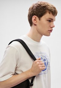 Camiseta blanca de algodón con un diseño gráfico azul en la parte delantera. El modelo sostiene una correa negra de una bolsa, con un fondo neutro.