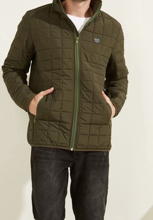 Homme portant une veste matelassée zippée vert olive avec les mains dans les poches et un jean foncé, debout devant un fond clair uni.