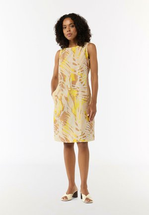Sleeveless dress in a yellow, beige, and brown leaf pattern; knee-length with side pockets; smooth fabric; paired with white heeled mules.
