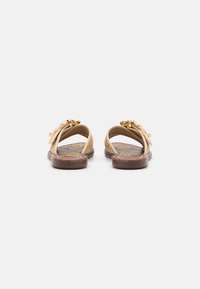 Sandales beiges avec une tige tressée et des éléments décoratifs en métal doré. Semelle plate marron avec des motifs texturés et un design à bout ouvert.