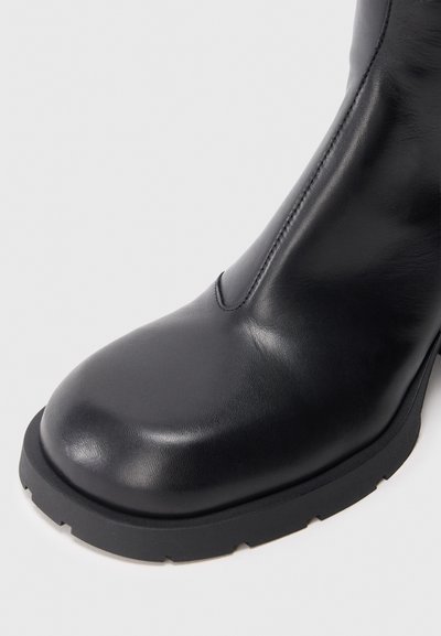 Bota de cano curto em couro preto com biqueira arredondada, textura lisa, detalhe de costura lateral e sola grossa preta em borracha com ranhuras para tração.