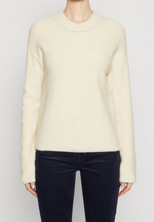 Personne portant un pull en maille côtelée crème à manches longues et un pantalon bleu marine foncé, posant devant un fond clair uni.
