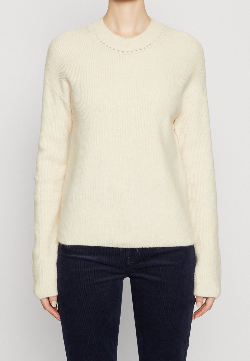 Personne portant un pull en maille côtelée crème à manches longues et un pantalon bleu marine foncé, posant devant un fond clair uni.