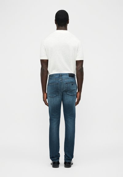 Άνδρας που φοράει λευκή κοντομάνικη πόλο μπλούζα και μπλε τζιν, στέκεται γυρισμένος με την πλάτη σε ένα απλό λευκό φόντο.