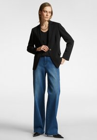 Blazer nero con un solo bottone, abbinato a jeans blu a gamba larga. Il blazer presenta un colletto a punta e tasche frontali.