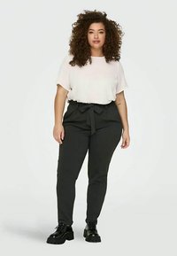Femme aux cheveux bouclés portant un haut blanc à manches courtes, un pantalon noir taille haute avec une ceinture nouée et des chaussures noires à semelle épaisse, se tenant devant un fond blanc.
